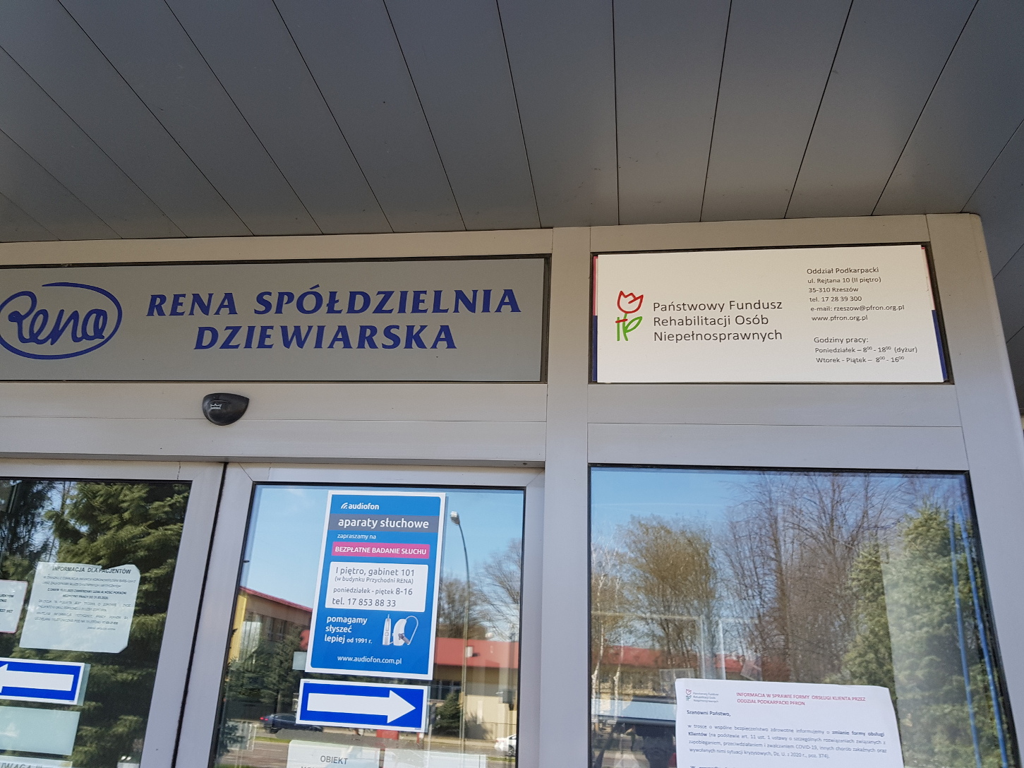 Wejście do budynku Wejście do budynku z drzwiami automatycznymi. całość wykonana z szarej stolarce aluminiowej. Na drzwiami logo Spółdzielni Dziewiarskiej „Rena”. Pop prawej stornie logo PFRON i informacje o godzinach działania Oddziału Podkarpackiego PFRON.