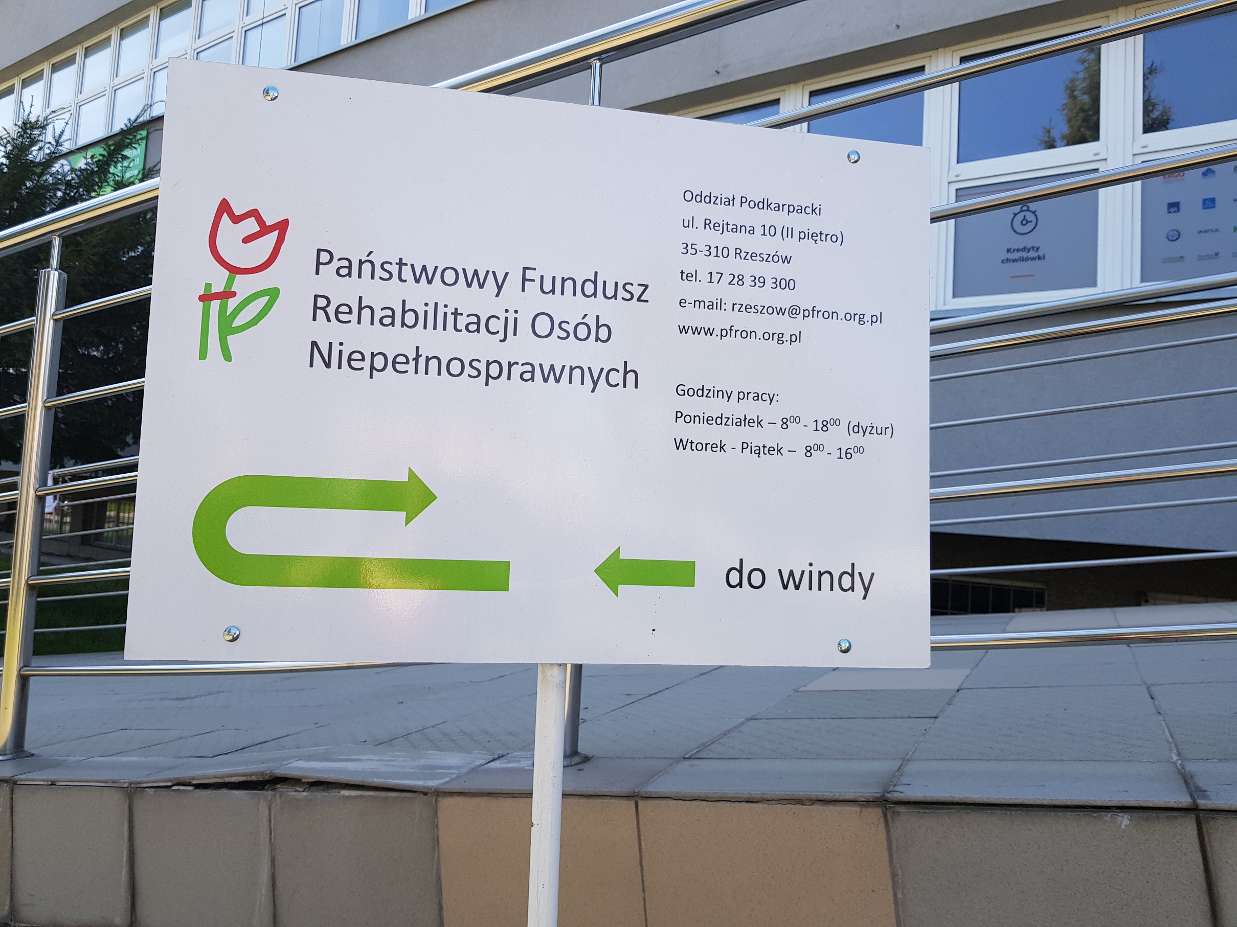 Tablica informacyjna Biała tablica informacyjna z logo PFRON i godzinami otwarcia Oddziału Podkarpackiego PFRON oraz drogowskazem, jaką droga dotrzeć do windy. W tle widać budynek biurowy.