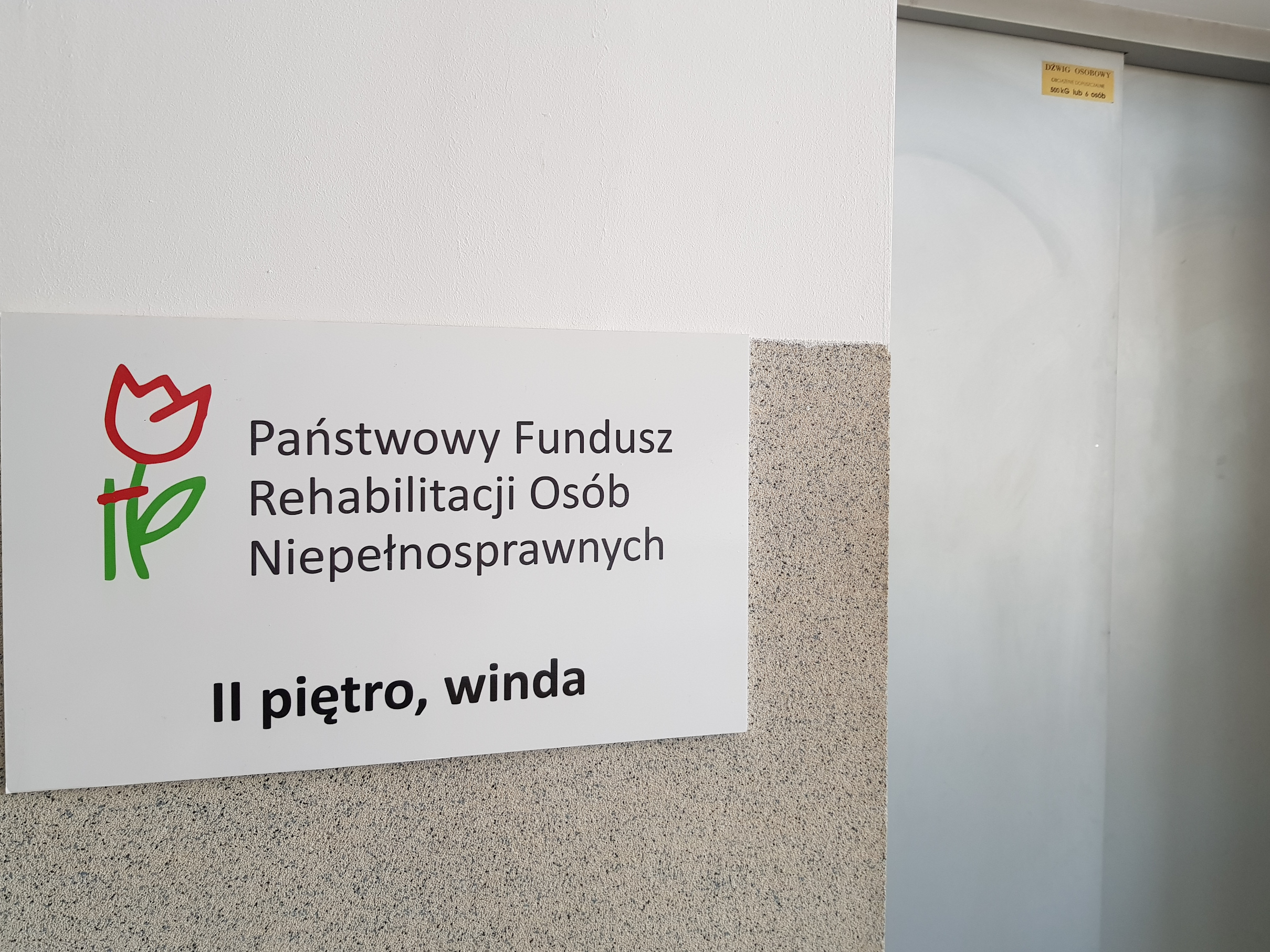 Tablica informacyjna przy windzie Na białej ścianie nad beżowymi lamperiami wisi tablica informacyjna z logo PFRON i informacją, gdzie mieści się Oddział - drugie piętro, dostępne za pomocą windy. Po prawej stronie od tablicy widoczne drzwi kabiny windy.