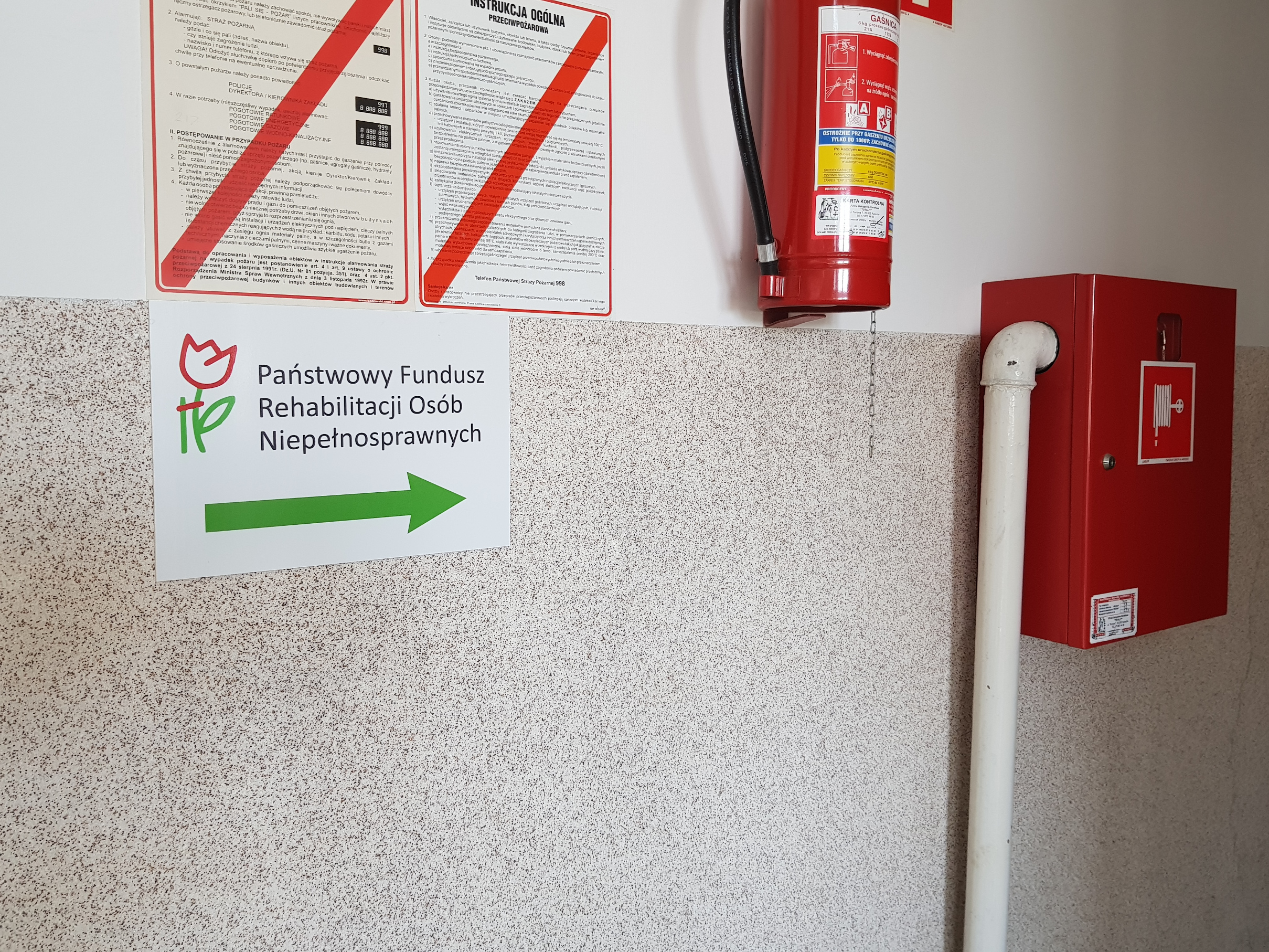 Tablica kierunkowa Biała ściana z beżową lamperią, nad którą wiszą instrukcje przeciwpożarowe. Po instrukcjami tablica z logo PFRON, wskazująca w którą stronę należy się kierować do Oddziału. Po [prawej stronie od instrukcji na ścianie wisi gaśnica , a poniżej niej widać skrzynkę hydrantu przeciwpożarowego.