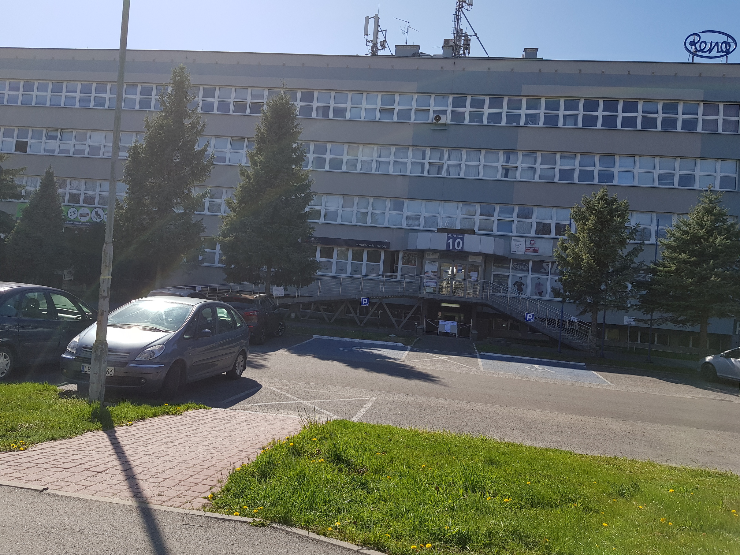 Budynek siedziby Oddziału Podkarpackiego PFRON - plan dalszy W tle widać czterokondygnacyjny budynek o szarej, mocno przeszklonej elewacji. Przed budynkiem widać drzewa oddzielające go od parkingu, na którym widoczne są dwa miejsca wyznaczone dla Osób niepełnosprawnych. Na parkingu widać tez zaparkowane samochody.