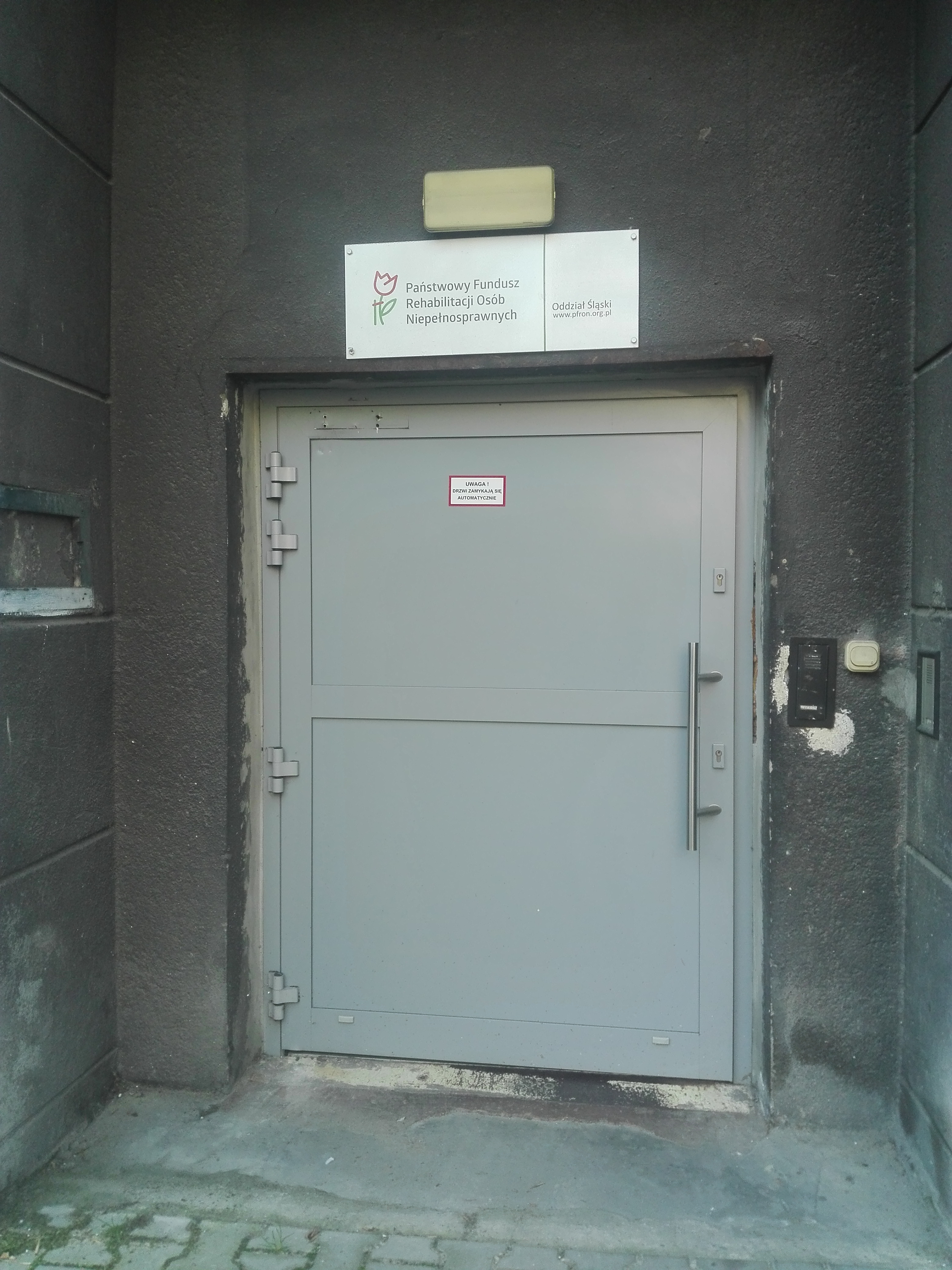 Wejście do Oddziału Śląskiego PFRON Szerokie jasnoszare drzwi. Nad drzwiami tablica z logo i napisem „Państwowy Fundusz Rehabilitacji Osób Niepełnosprawnych”. Po prawej stronie od drzwi dzwonek oraz domofon. Na drzwiach tabliczka z napisem „uwaga, drzwi zamykają się automatycznie”