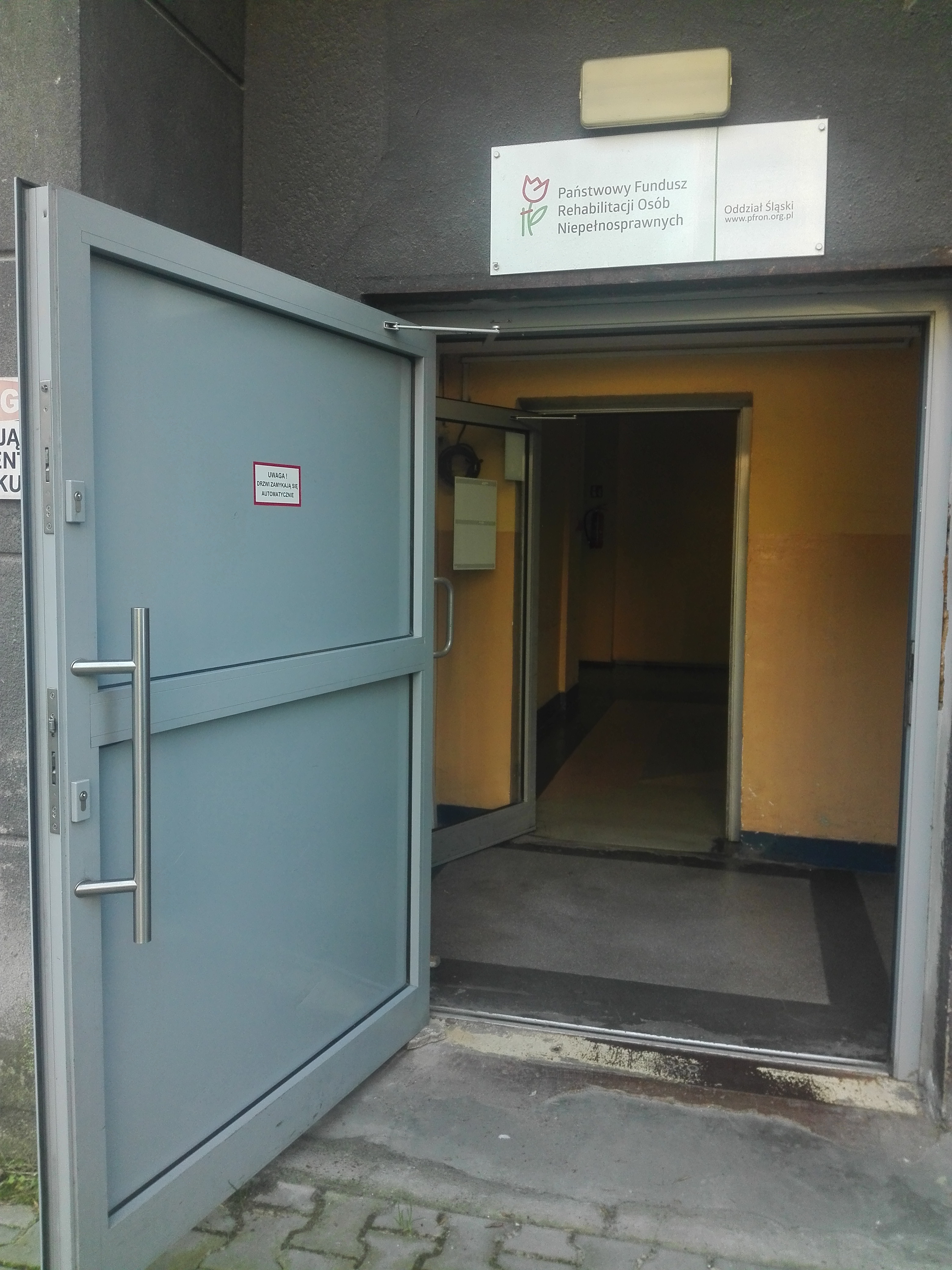 Wejście do Oddziału Śląskiego PFRON - otwarte Otwarte drzwi do budynku. Na drzwiach tabliczka z napisem „uwaga, drzwi zamykają się automatycznie”. Za nimi przedsionek gdzie mamy drugie - przeszklone i jednoskrzydłowe drzwi, otwarte i prowadzące w głąb budynku.