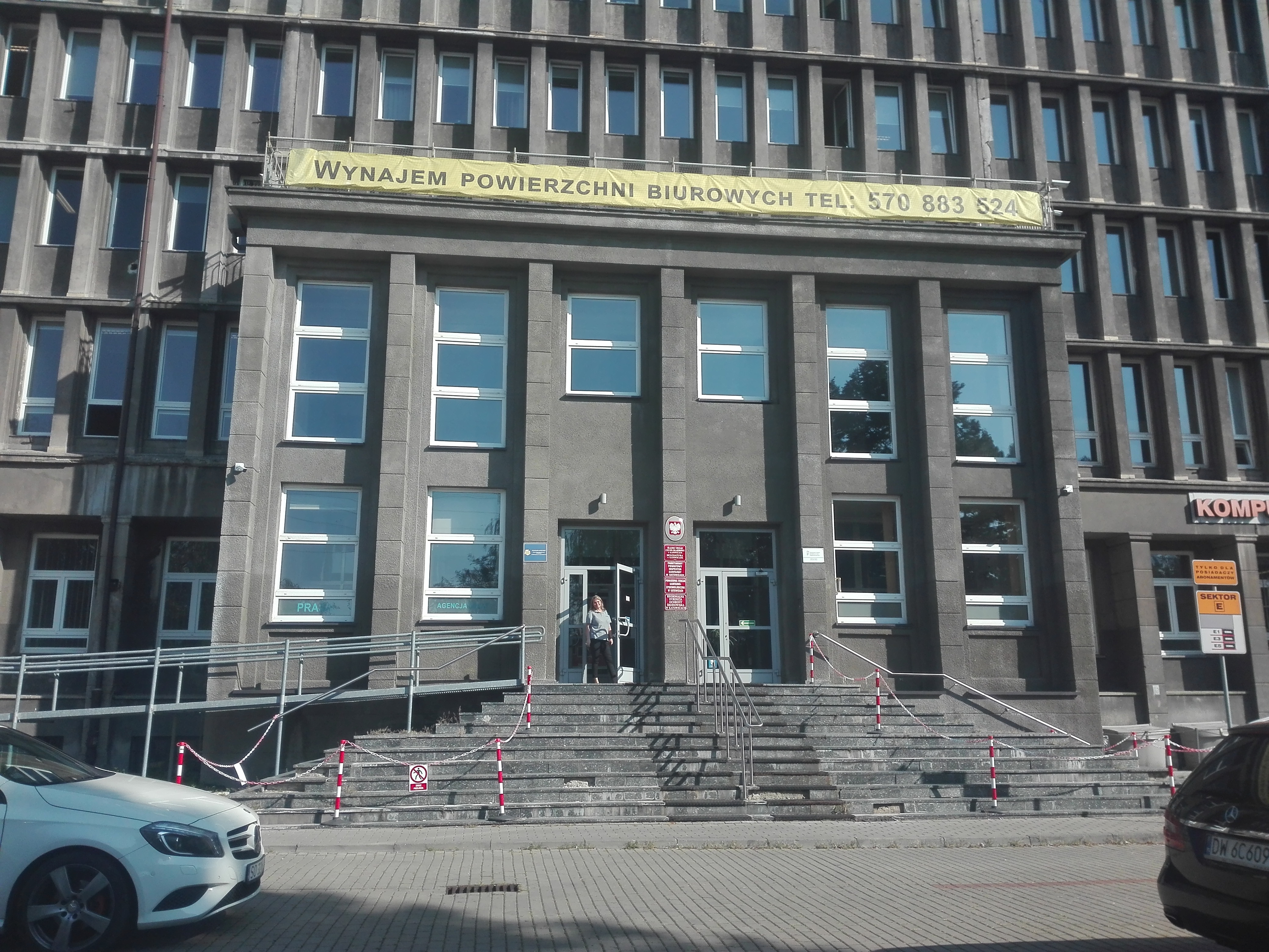 Siedziba oddziału Śląskiego PFRON Budynek biurowy - główne wejście, do którego prowadzą schody. Po lewej stronie schodów pochylnia dla wózków. Na schodach wyznaczone ciągi komunikacyjne i miejsca, gdzie zakazano ruchu pieszego. Przed wejściem na ulicy zaparkowane samochody. Nad wejściem baner reklamowy „Wynajem powierzchni biurowych” wraz z numerem telefonu