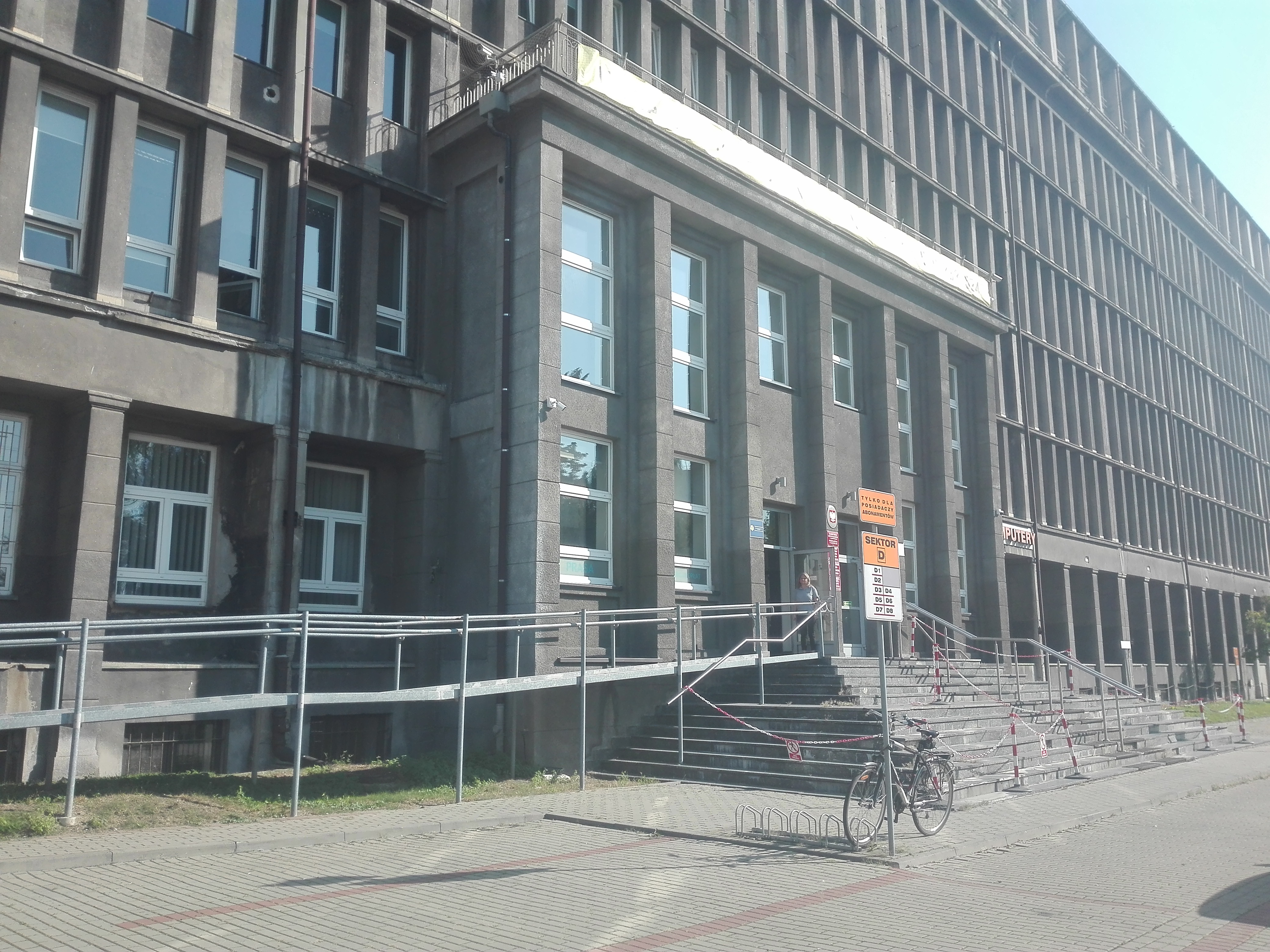 Wejście do siedziby Oddziału Śląskiego PFRON - ujęcie z parkingu Główne wejście do budynku biurowego, na pierwszym planie parking dla rowerów z jednym zaparkowanym rowerem. Po lewej stronie od wejścia widzimy pochylnię dla wózków.