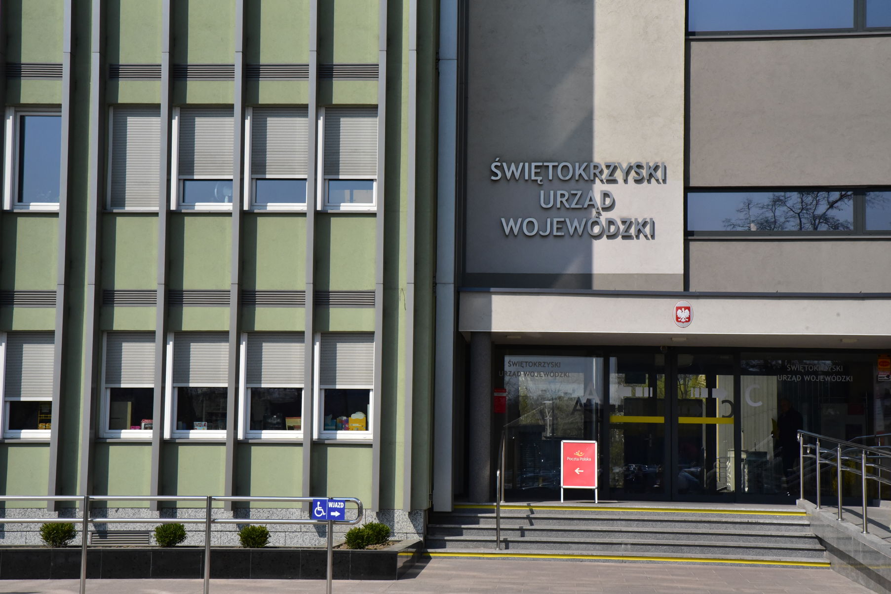 Wejście od Oddziału Świętokrzyskiego PFRON Budynek o zielonej elewacji z szarymi elementami. Po prawej stronie schody wejściowe do budynku, prowadzące do szklanych drzwi, nad drzwiami daszek, nad którym umieszczone napis "Świętokrzyski Urząd Wojewódzki". Po lewej storni widać poręcz, na której zainstalowany jest znak z napisem "wjazd" i piktogramem osoby niepełnosprawnej prawej stronie do schodów widać pochylnię prowadzącą do wejścia.