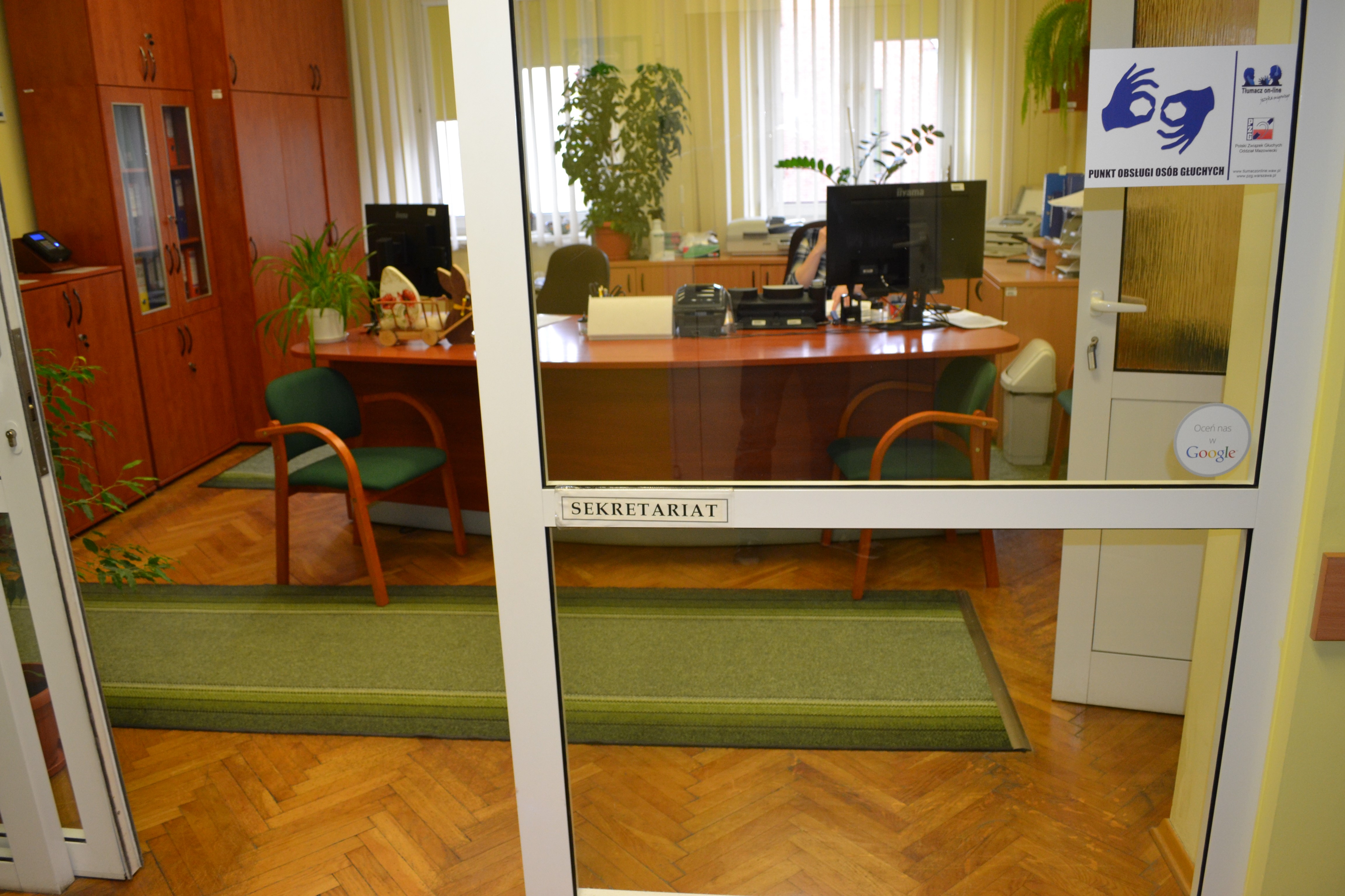 Widok sekretariatu. Na pierwszym planie widać szklaną ścianę, na której zamocowano poprzeczkę, na której z kolei powieszono tabliczkę „sekretariat”. Widoczne wnętrze pomieszczenia. Na pierwszym polanie zielony dywanik, za którym stoi duże biurko, zastawione sprzętem biurowym. Po lewej stronie od biurka szafa, a po prawej otwarte drzwi do dalszego pomieszczenia.