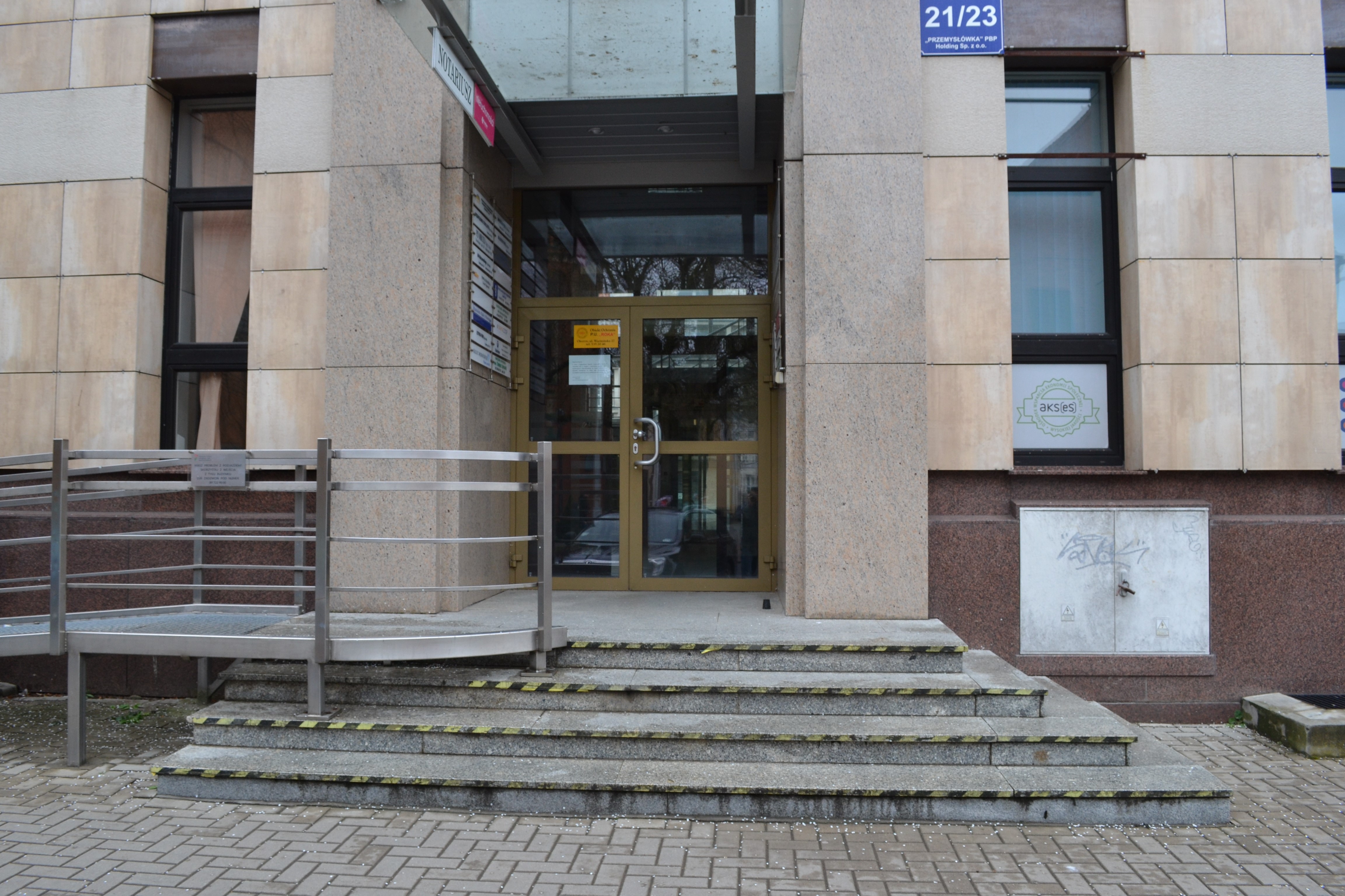 Główne drzwi wejściowe do budynku. Podwójne, przeszklone drzwi, schowane pod szklanym daszkiem. Przed drzwiami podest, do którego prowadzą schody, skontrastowane na krawędziach. Z prawej strony na podest prowadzi pochylnia dla wózków.