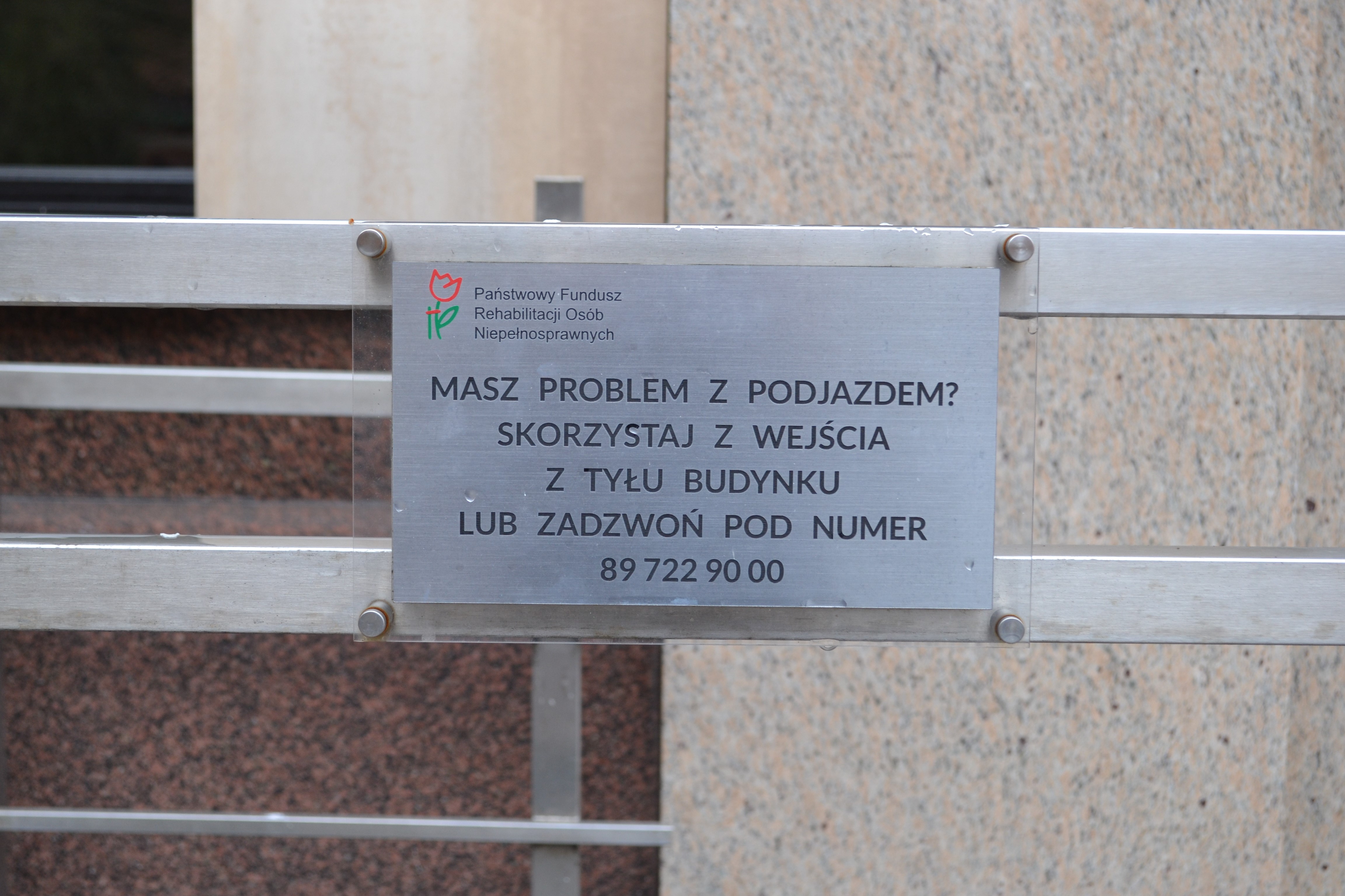 Tabliczka na pochylni dla wózków prowadzącej do głównego wejścia budynku. Tekst na tabliczce brzmi: „Masz problem z podjazdem? Skorzystaj z wejścia z tyłu budynku lub zadzwoń pod numer 89 722 90 00”. Nad napisem, w lewym górnym rogu tabliczki logo PFRON i napis „Państwowy Fundusz Rehabilitacji Osób Niepełnosprawnych”
