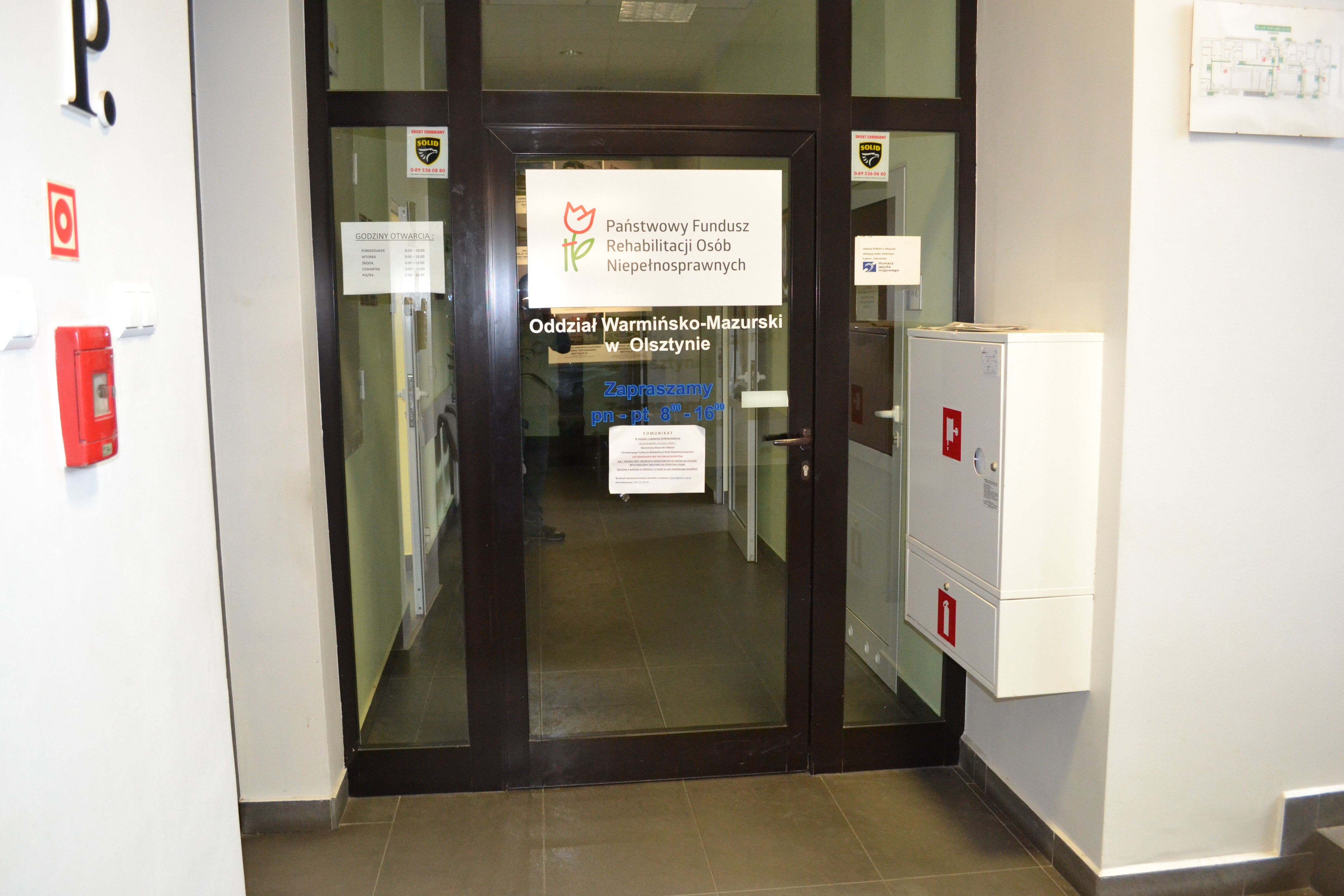 Drzwi wejściowe do Oddziału. Pośrodku drzwi duża biała tablica z logo PFRON i napisem „Państwowy Fundusz Rehabilitacji Osób Niepełnosprawnych”. Pod tablicą nazwa Oddziału i godziny urzędowania. Na prawo do drzwi, na ścianie białe skrzynki z hydrantem przeciwpożarowym
