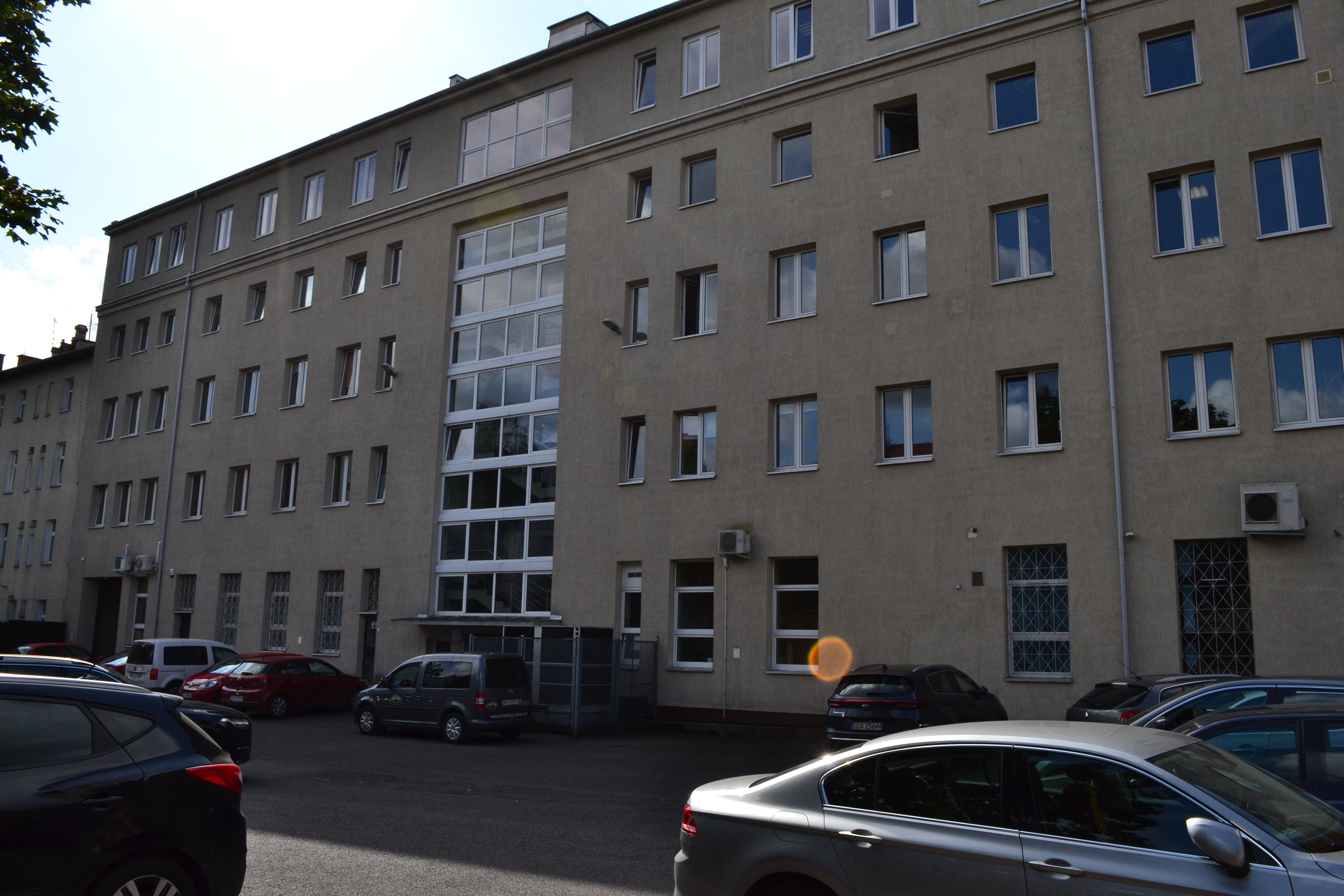 Widok budynku od strony dziedzińca. Liczne zaparkowane samochody. Na ścianie, przy niektórych oknach wiszą klimatyzatory.