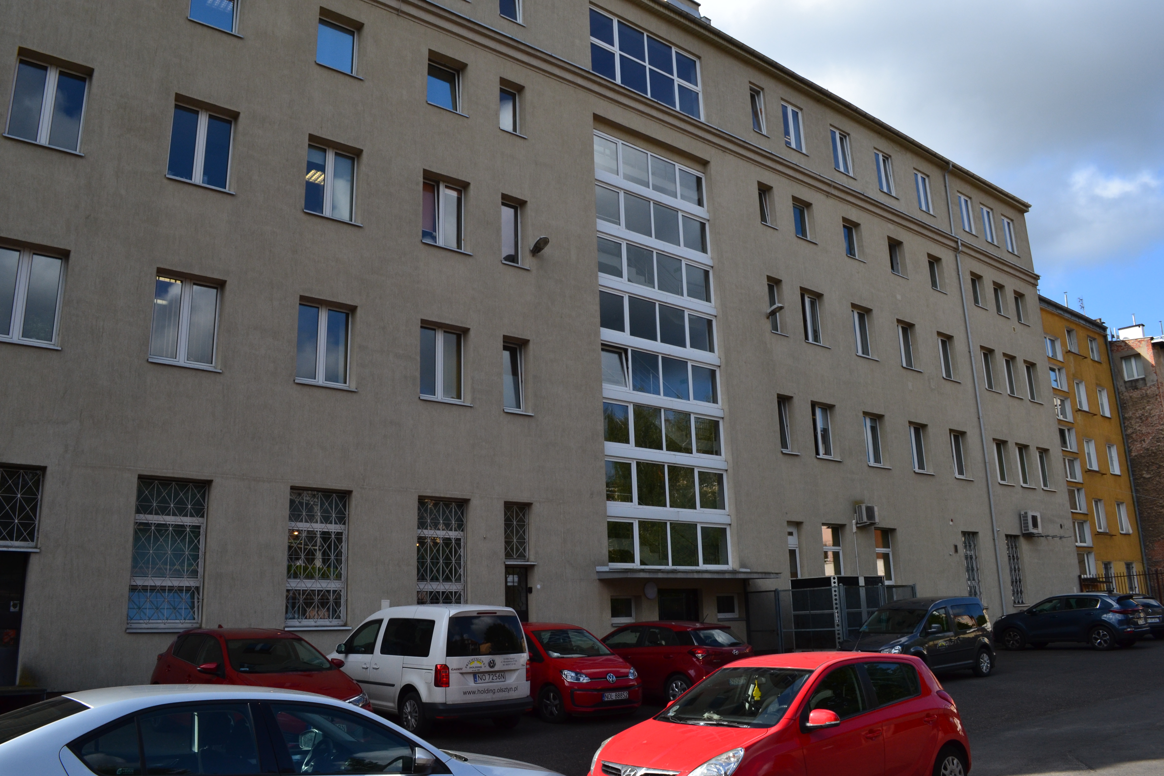 Budynek od strony dziedzińca. Widoczne liczne samochody , zaparkowane na dziedzińcu. Na budynku doskonale widoczna klatka schodowa, wyróżniając się dużym przeszkleniem. Na ścianie, przy niektórych oknach wiszące klimatyzatory.