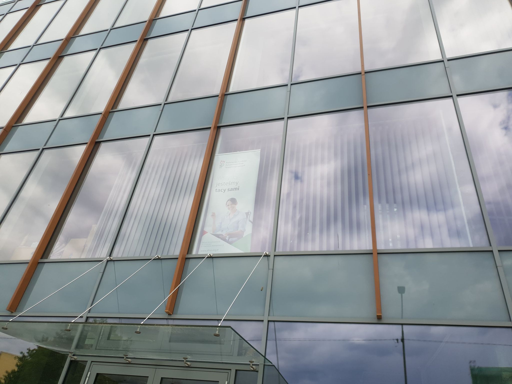 Elewacja budynku Szklana elewacja budynku. Przez przezroczystą szybę widać duży roll-up z sylwetką kobiety i logiem Państwowego Funduszu Rehabilitacji Osób Niepełnosprawnych.