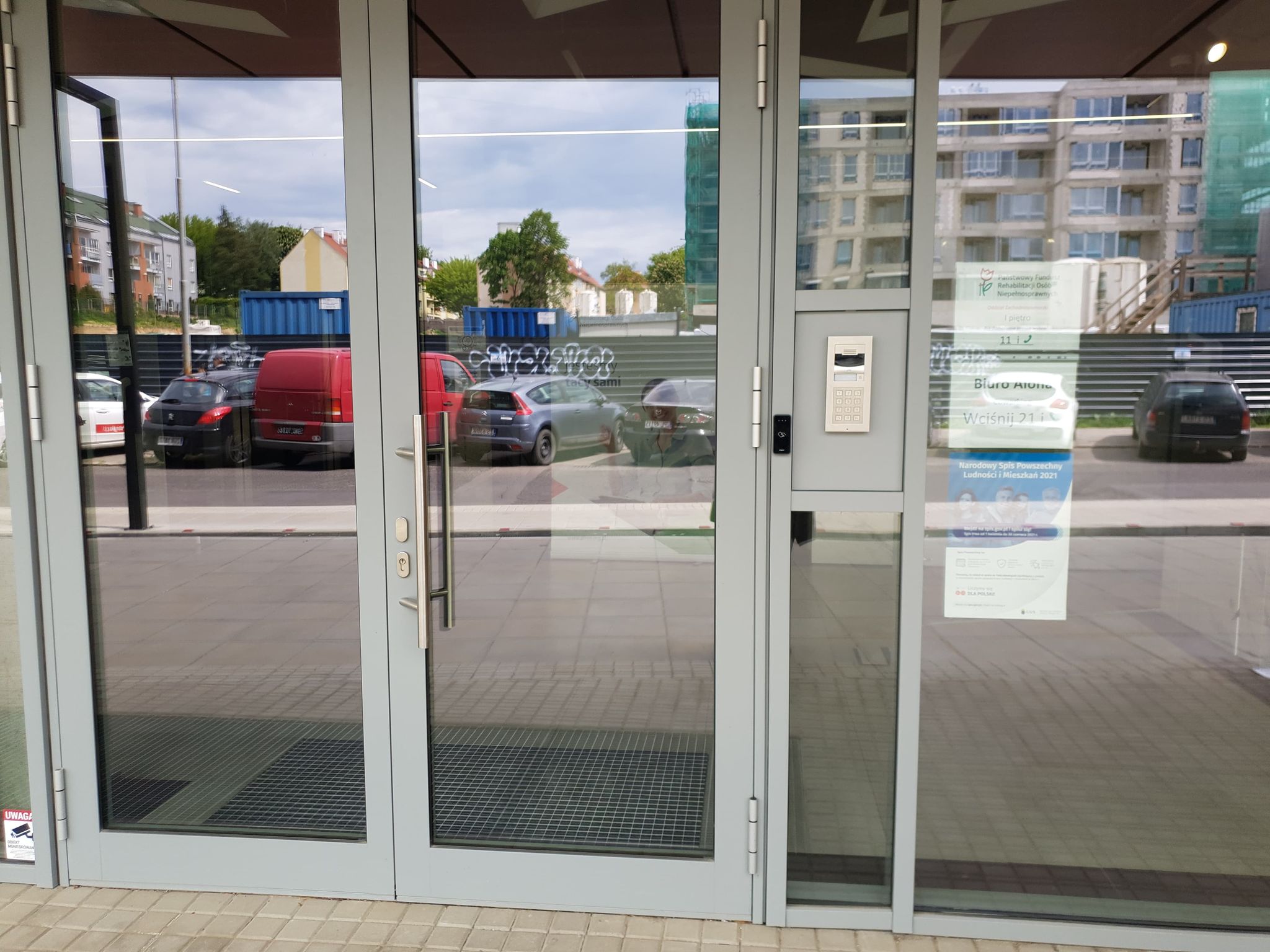 Wejście do budynku Drzwi wejściowe do budynku, dwuskrzydłowe. Po prawej stronie od drzwi domofon. Obok domofonu tabliczki informacyjne o instytucjach znajdujących się w budynku> Na samej górze logo Państwowego Funduszu Rehabilitacji Osób Niepełnosprawnych. W drzwiach i szklanej ścianie odbija się sąsiadujące otoczenie