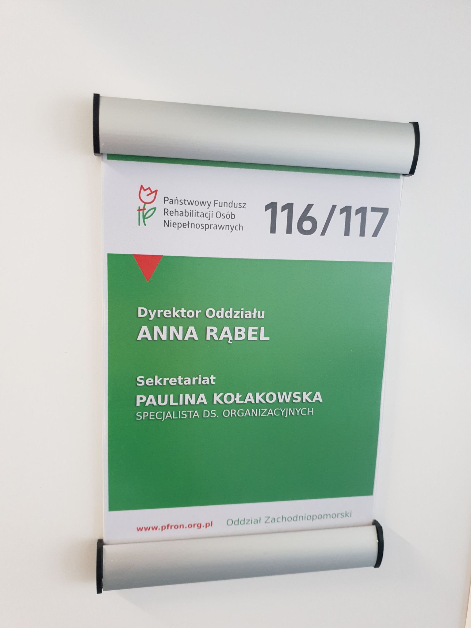 Tabliczka informacyjna Tabliczka informacyjna z oznaczeniem pomieszczenia o numerze 116/ 117. Na tabliczce, na zielonym tle umieszczono imiona i nazwiska osób, pracującyh w tych pomieszczeniach: Dyrektor Oddziału oraz Specjalista ds. organizacyjnych. Powyżej te informacji logo i napis Państwowy Fundusz Rehabilitacji Osób Niepełnosprawnych. Pod danymi na samym dole tabliczki adres internetowy PFRON.
