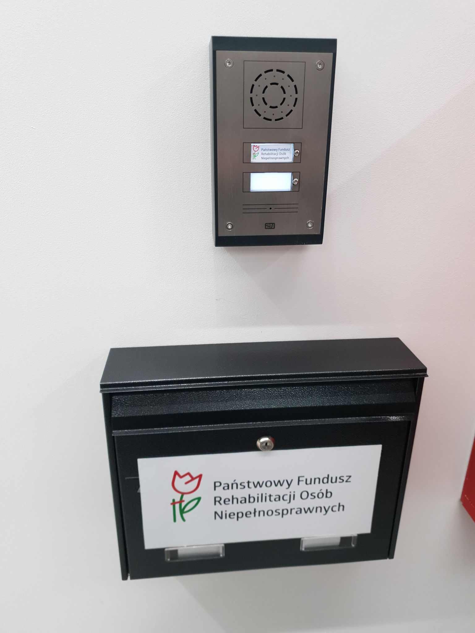 Domofon i skrzynka podawcza Domofon w kolorze metalu, umieszczony centralnie na białej scianie, z dużym przyciskiem, opisany logiem i napisem „Państwowy Fundusz Rehabilitacji Osób Niepełnosprawnych”. Pod domofonem umieszczona czarna skrzynka na dokumenty, również oznaczona w taki sam sposób