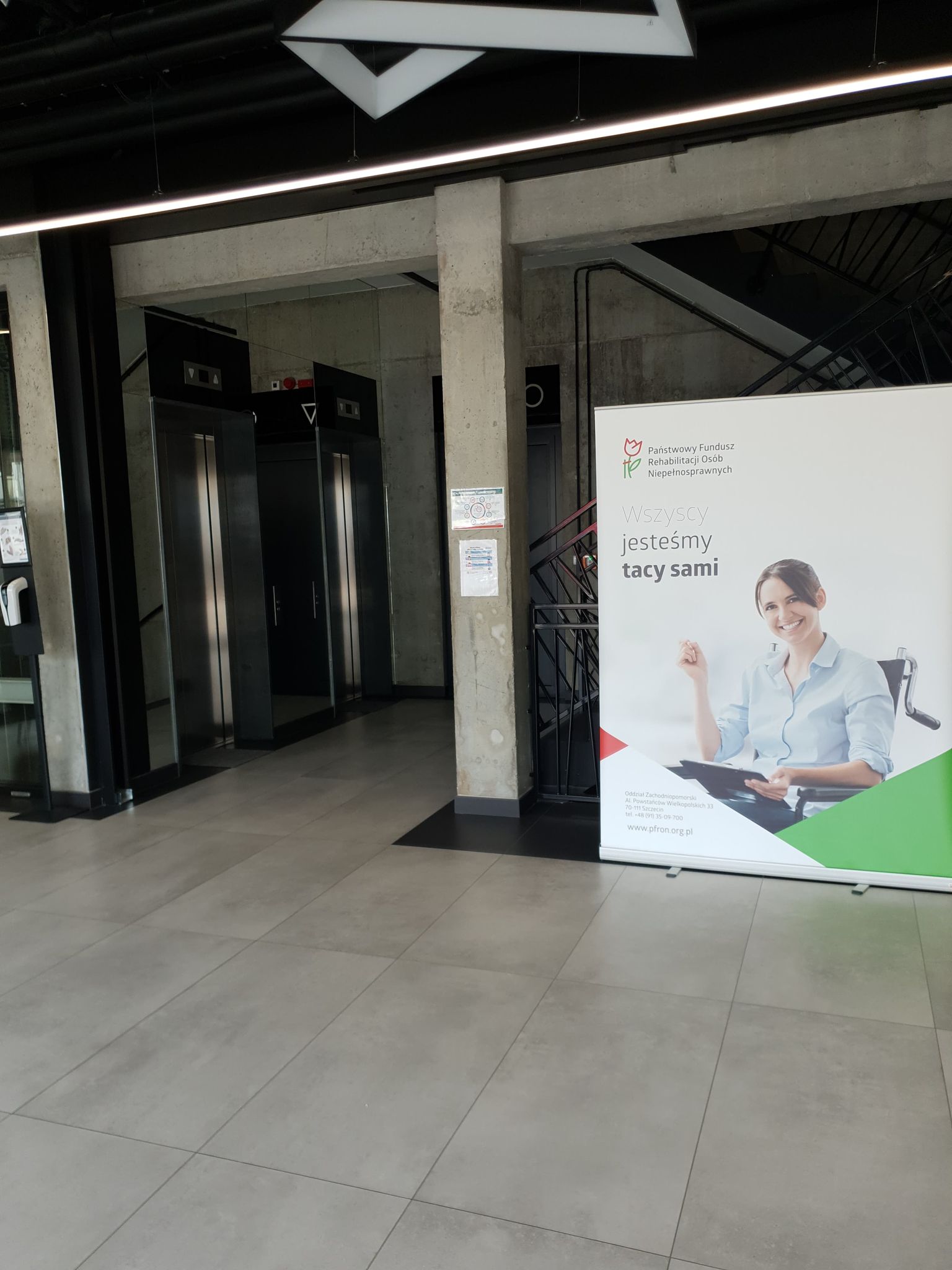 Hol na parterze Hol wewnętrzny budynku, w tle widać szyby wind, a po prawej stronie od nich schody przesłonięte dużym roll-upem z reklamą Państwowego Funduszu Rehabilitacji Osób Niepełnosprawnych.